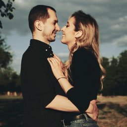 RUSLAN BURMISTROV - Zakochana para w czarnych strojach, uśmiechnięta, obejmująca się na tle pochmurnego nieba i drzew podczas sesji plenerowej.