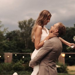 RUSLAN BURMISTROV - Uśmiechnięta panna młoda w białej sukni, trzymana w objęciach przez pana młodego w brązowym garniturze, z bukietem ślubnym w ręku, na tle zieleni i dekoracyjnych światełek.