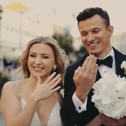 RUSLAN BURMISTROV - Uśmiechnięta para młoda prezentuje obrączki na tle miejskiej ulicy. Panna młoda w sukni z delikatnymi ramiączkami, pan młody w czarnym smokingu z białą butonierką i bukietem białych kwiatów.