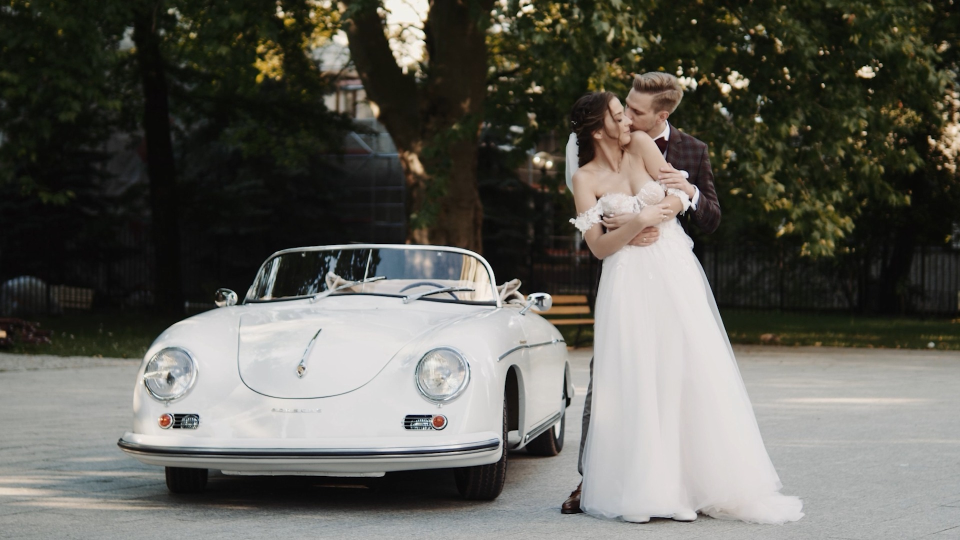 Para młoda w objęciach przed białym, klasycznym Porsche 356 Speedster na tle zieleni drzew