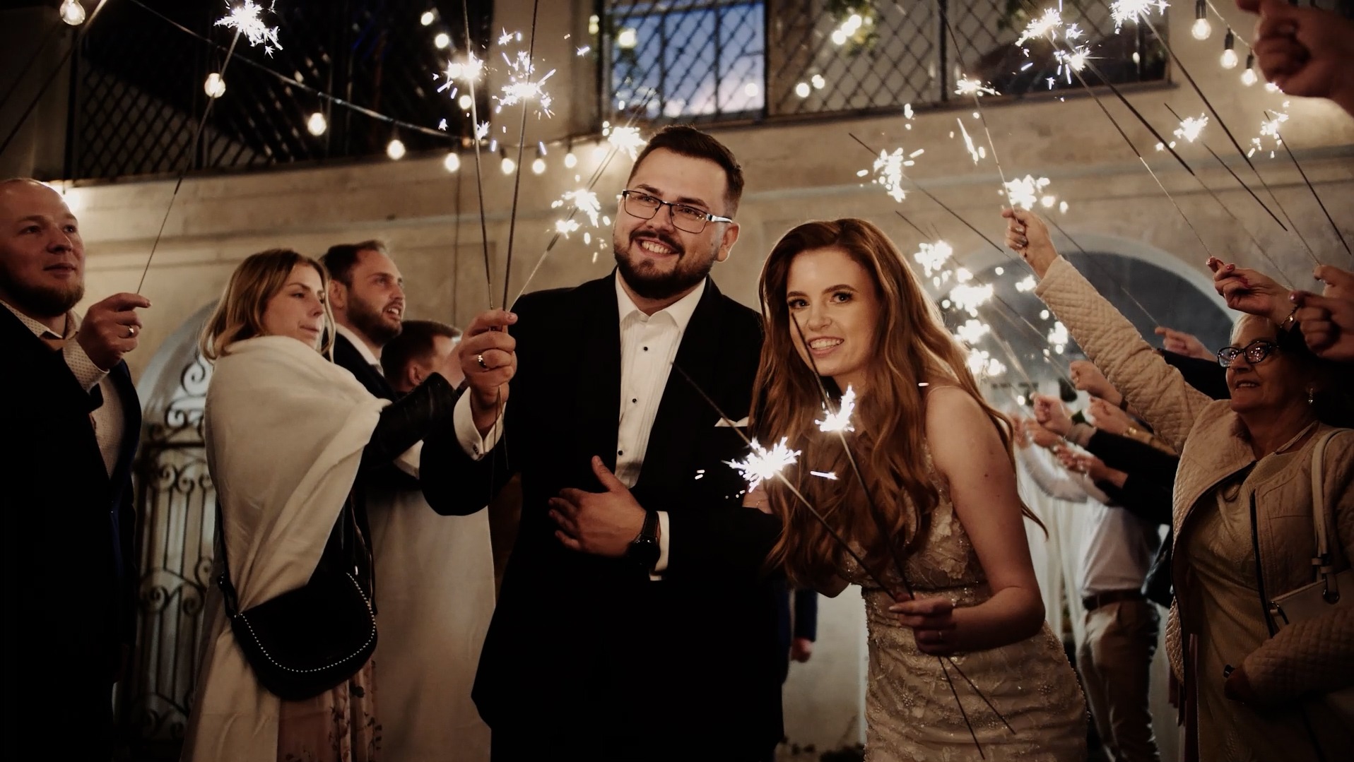 Uśmiechnięta para młoda otoczona gośćmi z palącymi się zimnymi ogniami podczas wieczornego przyjęcia weselnego, oświetlona dekoracyjnymi żarówkami.