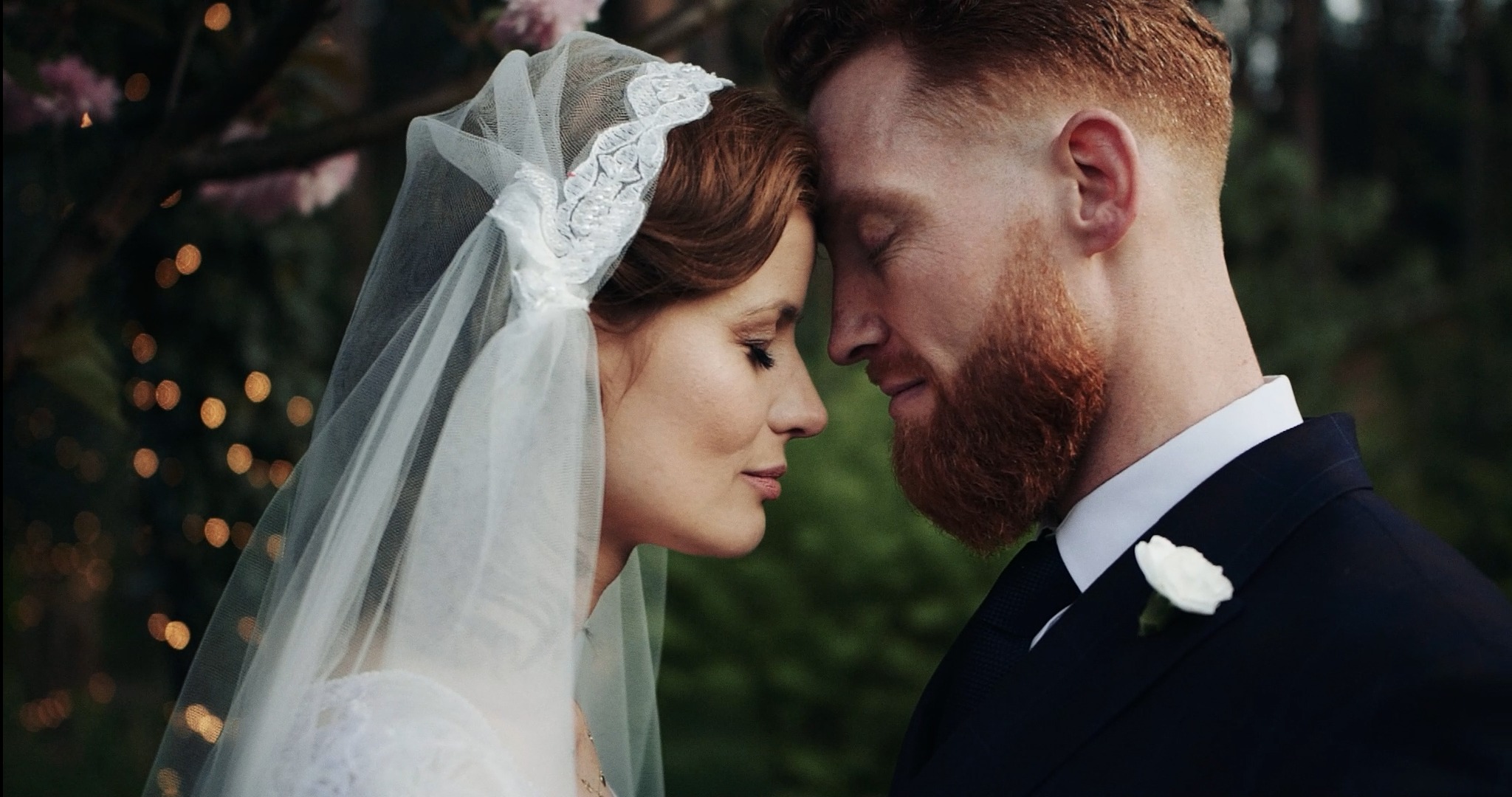 Zbliżenie na parę młodą, stykającą się czołami, panna młoda w białej sukni i welonie z koronkowym wykończeniem, pan młody z rudą brodą w ciemnym garniturze z białą butonierką, w tle rozmyte światełka.