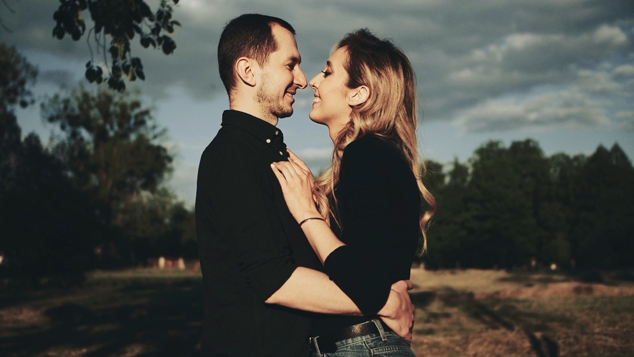 Zakochana para w czarnych strojach, uśmiechnięta, obejmująca się na tle pochmurnego nieba i drzew podczas sesji plenerowej.