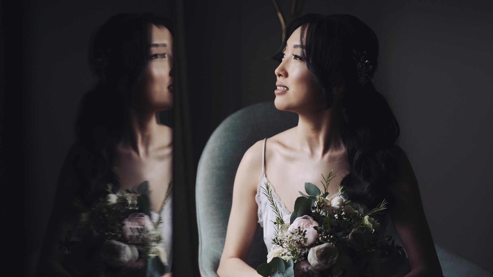 Panna młoda w jasnej sukience siedzi na krześle z bukietem, odbicie w lustrze, delikatne oświetlenie.