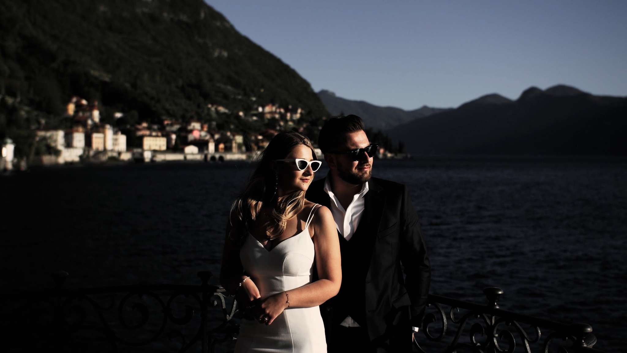 Elegancka para w okularach przeciwsłonecznych pozuje na tle jeziora i gór, prawdopodobnie podczas sesji ślubnej.