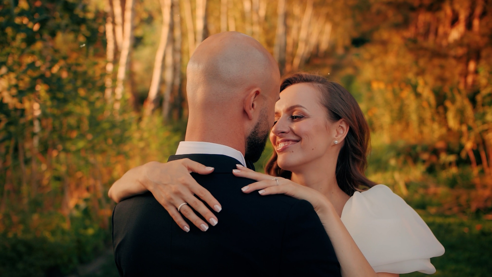 Uśmiechnięta panna młoda w objęciach pana młodego, jesienne tło z drzewami, ciepłe oświetlenie.