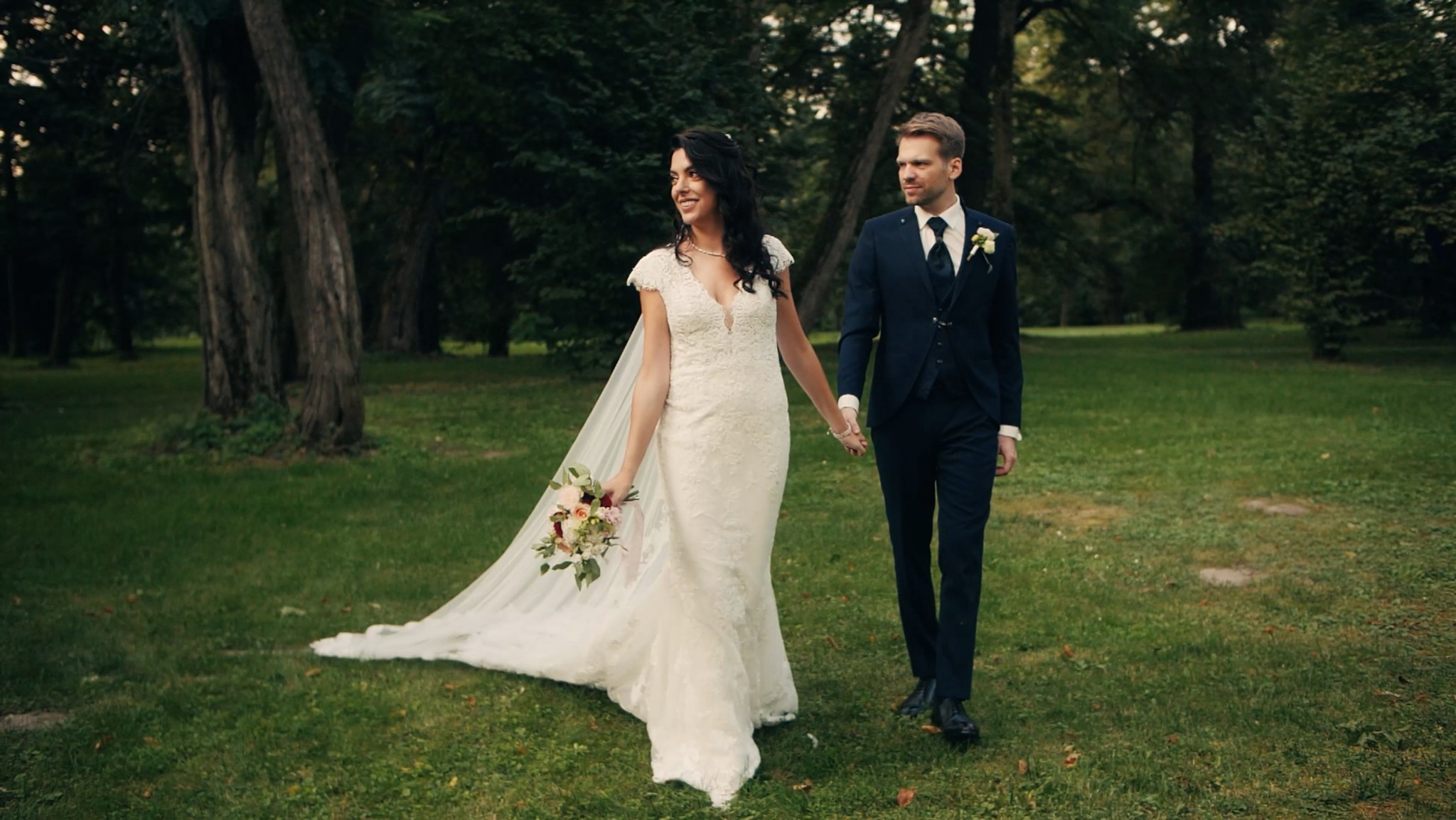 Młoda para w strojach ślubnych spaceruje po zielonym parku, trzymając się za ręce; panna młoda ma bukiet kwiatów.