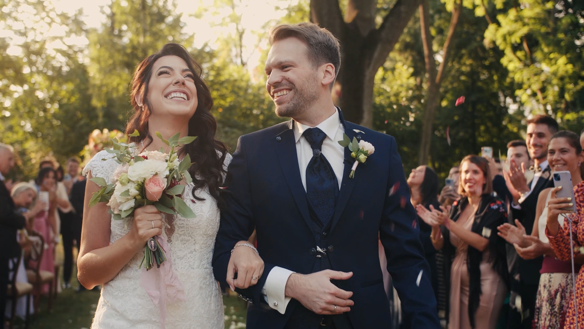 Uśmiechnięta para młoda, idąca alejką w otoczeniu gości, z konfetti opadającym z góry podczas ceremonii ślubnej w plenerze.