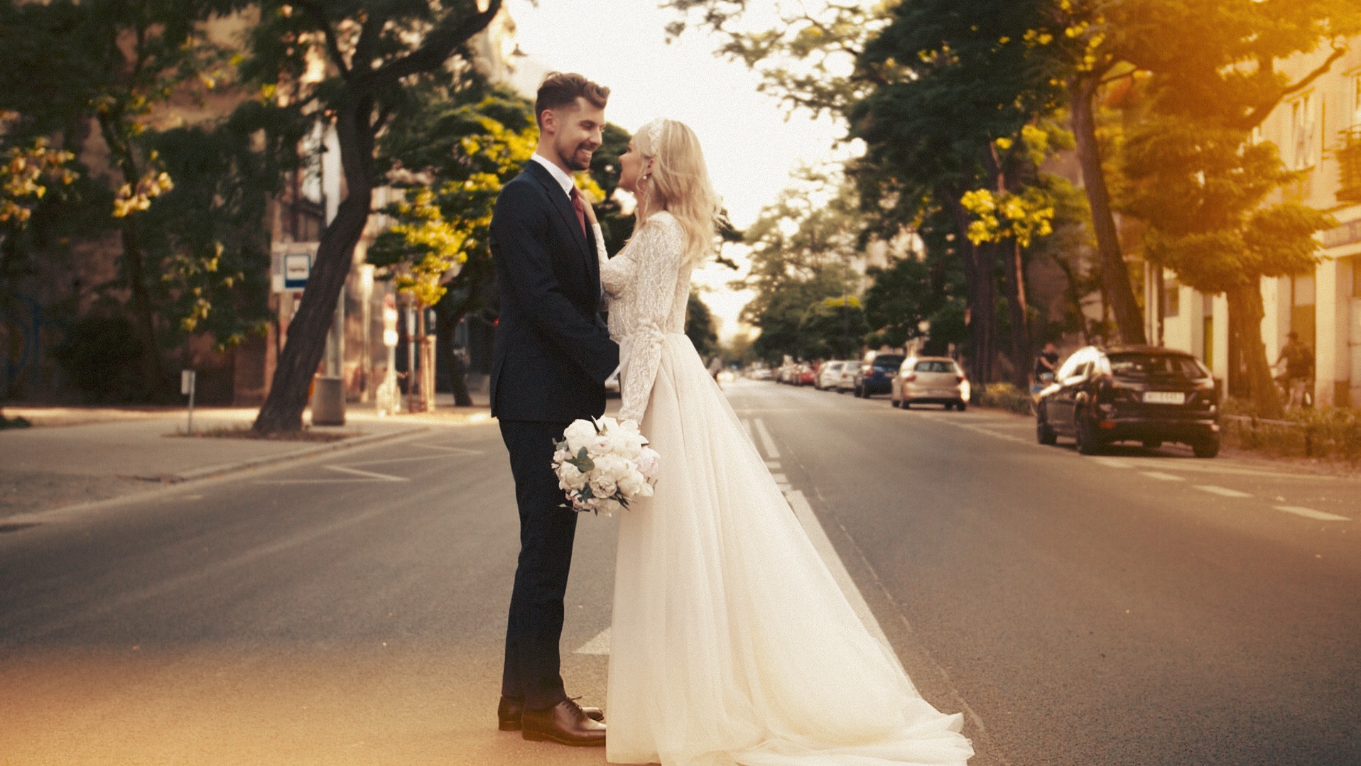 Para młoda stoi na pasach na ulicy, panna młoda w koronkowej sukni trzyma bukiet, w tle drzewa i budynki w ciepłym świetle.