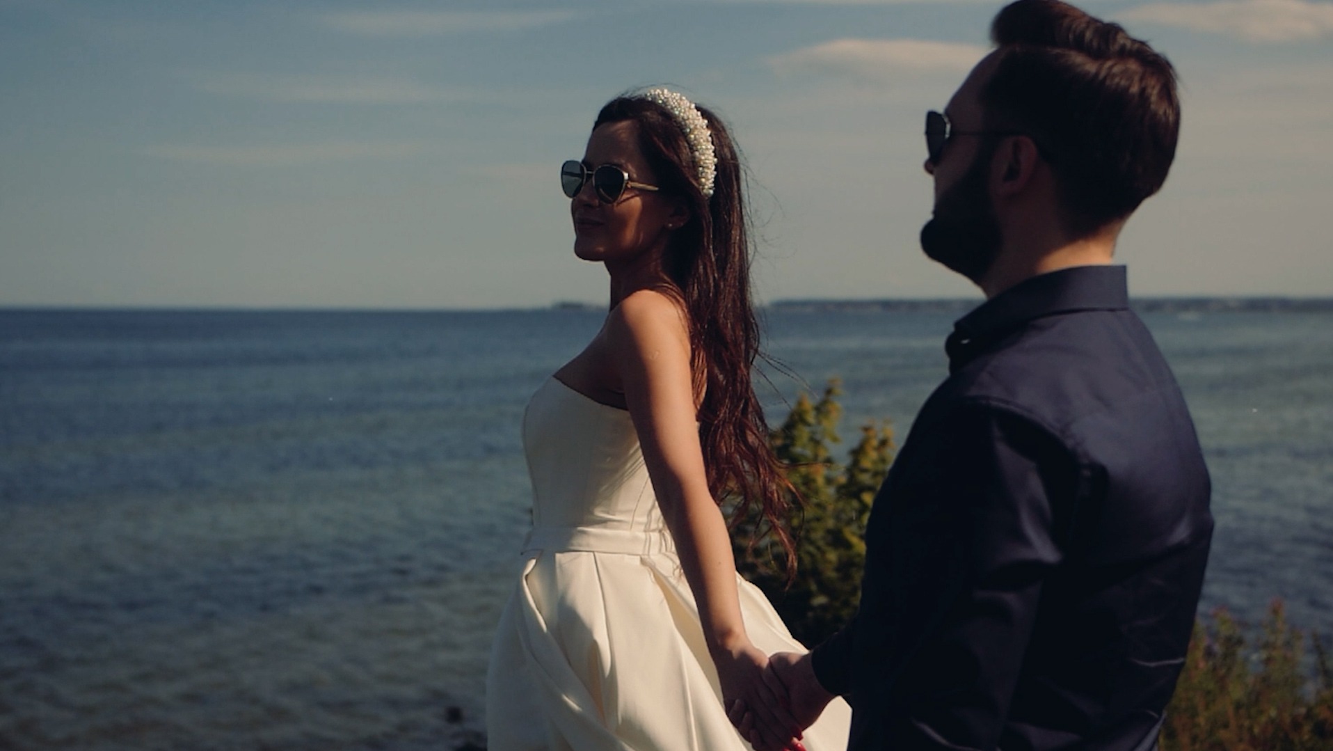 Para młoda w okularach przeciwsłonecznych, trzymająca się za ręce na tle morza w słoneczny dzień. Panna młoda w białej sukni bez ramiączek i z perłową opaską.