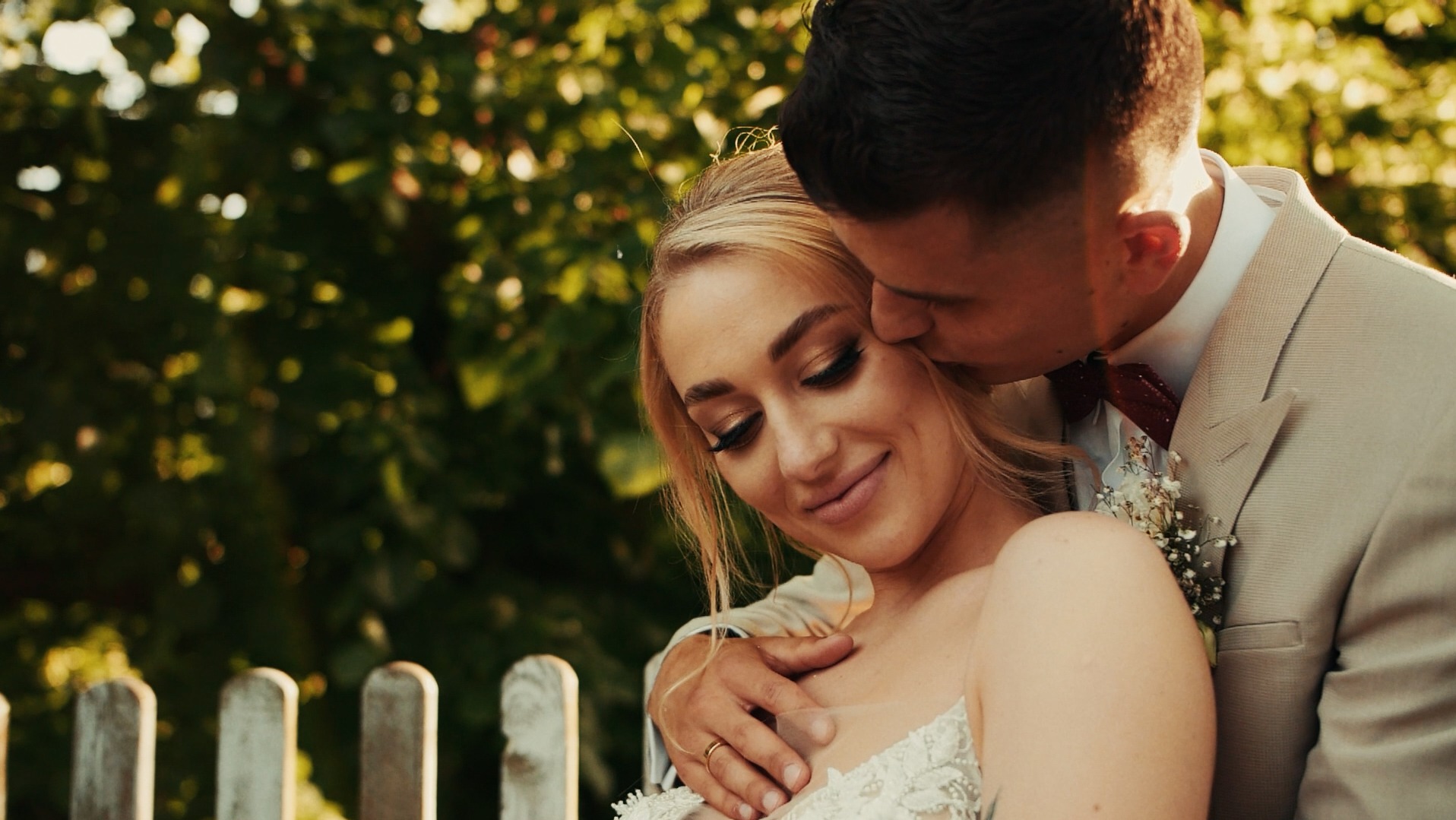 Panna młoda w koronkowej sukni uśmiecha się, gdy pan młody w beżowym garniturze i bordowej muszce całuje ją w policzek na tle zieleni i białego płotu.