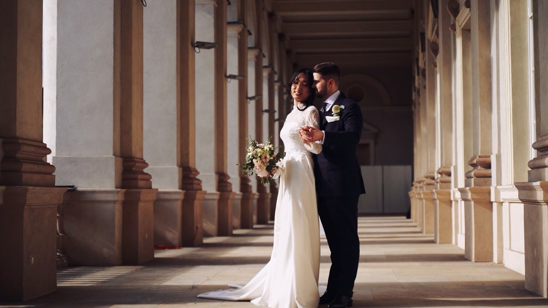 Para młoda w objęciach, panna młoda trzyma bukiet na tle kolumnady w ciepłym świetle