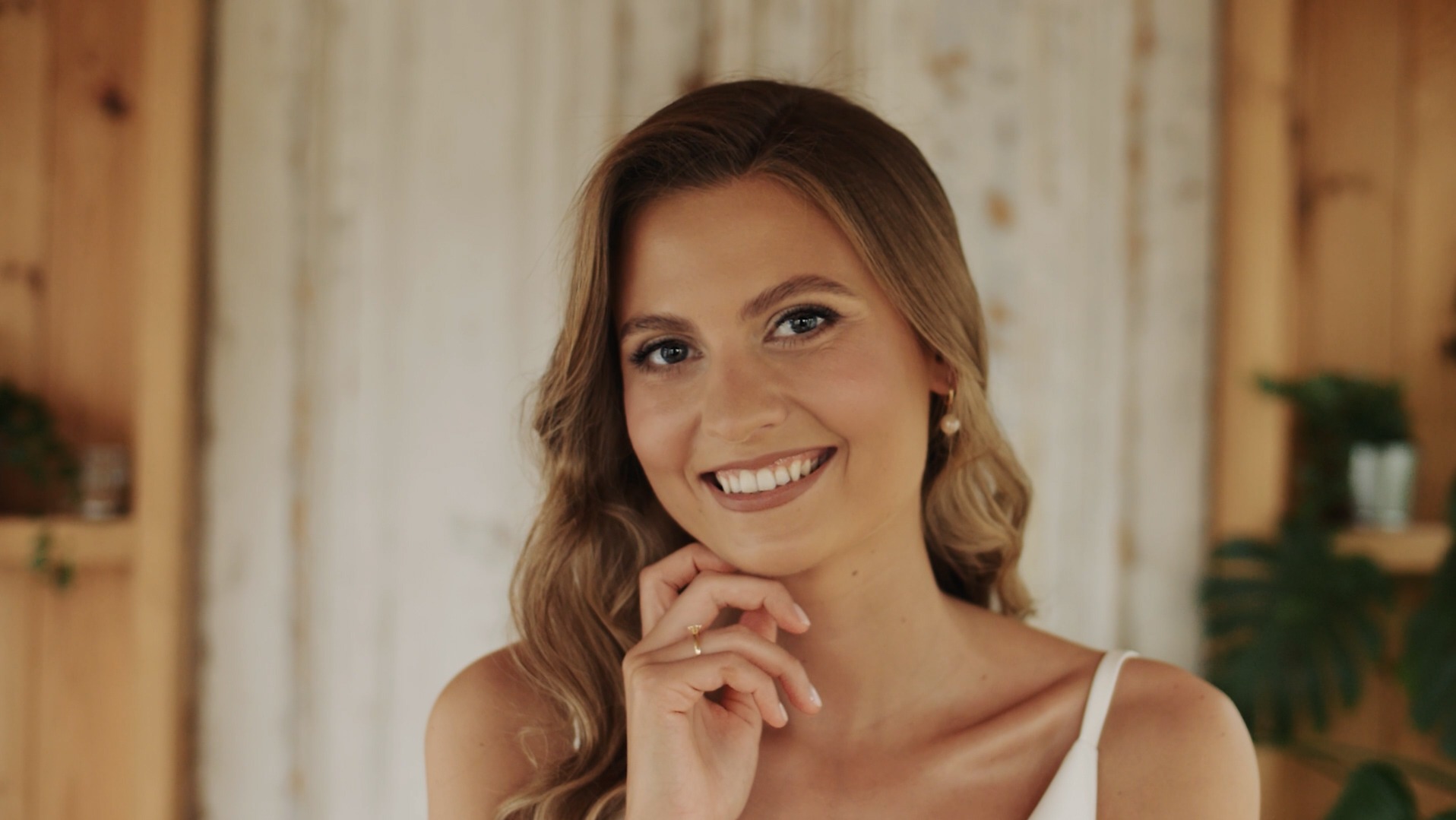 Portret uśmiechniętej kobiety z długimi blond włosami, delikatnym makijażem i złotą biżuterią, pozującej na tle drewnianego wnętrza z roślinami.