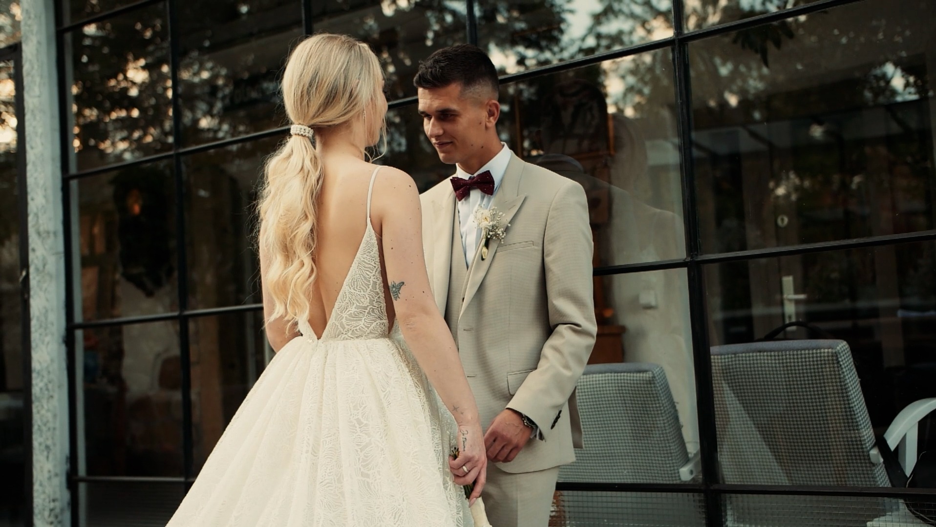 Młoda para w dniu ślubu: Panna Młoda w sukni z odkrytymi plecami i tatuażem motyla, Pan Młody w beżowym garniturze i bordowej muszce, w tle okno z odbiciem wnętrza.