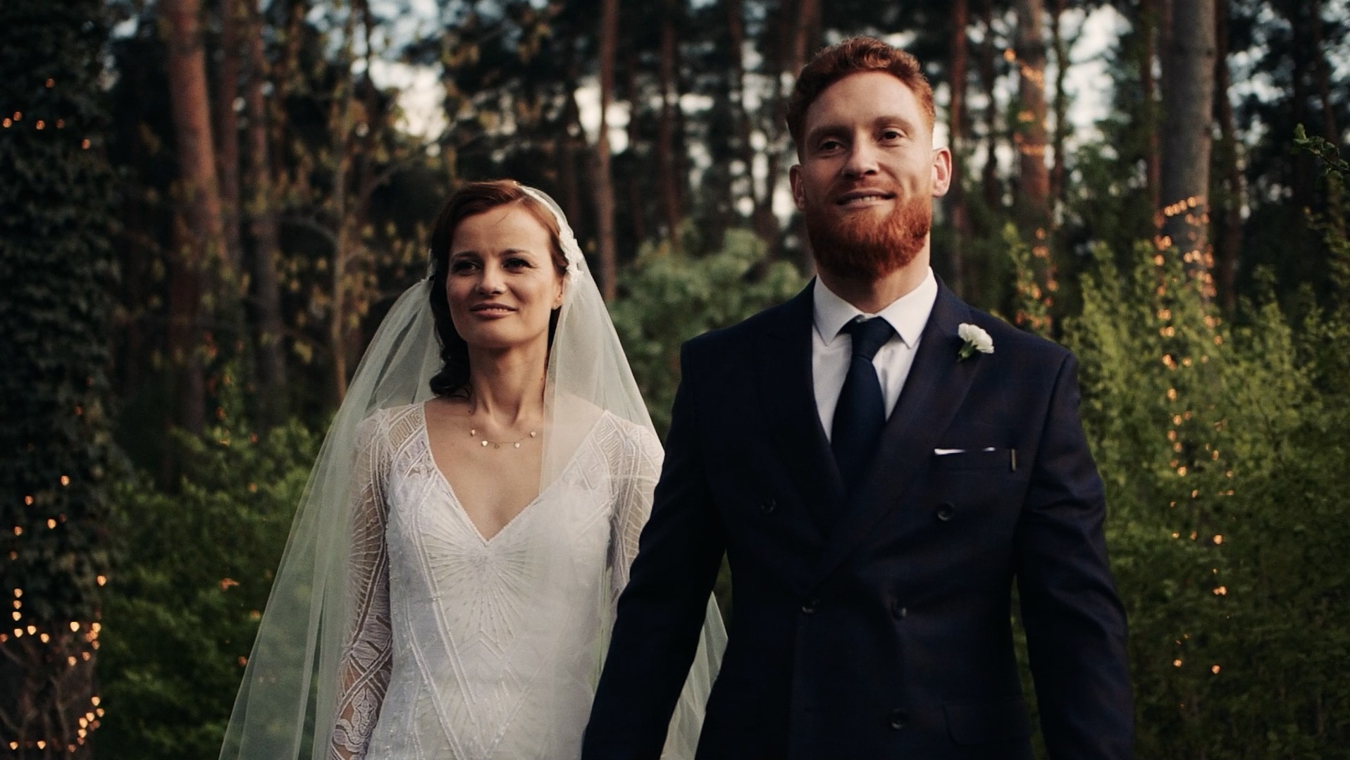 Para młoda w leśnej scenerii; panna młoda w sukni z koronkowymi rękawami, pan młody w granatowym garniturze z butonierką, delikatne oświetlenie w tle.