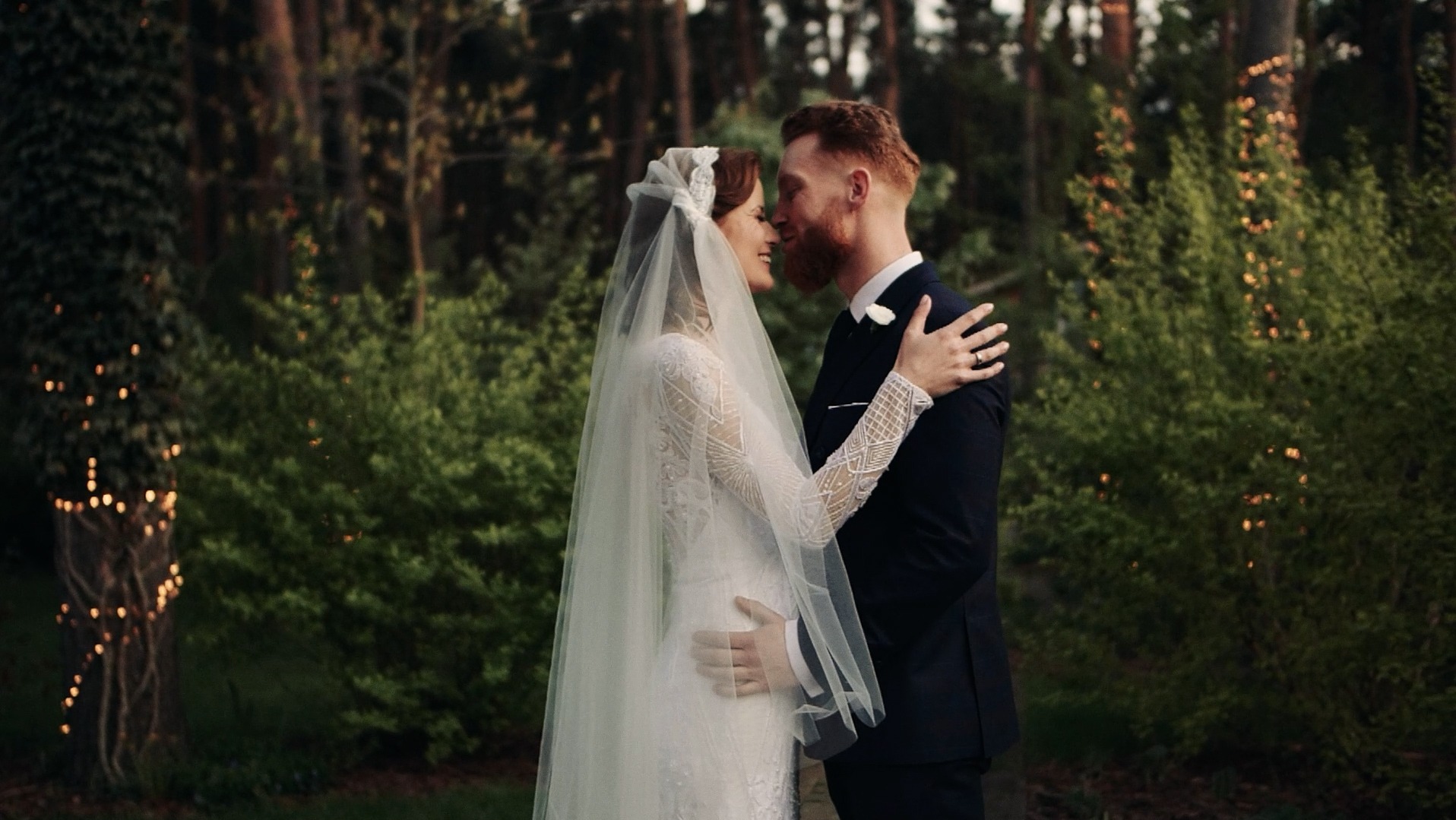Ujęcie profilowe pary młodej podczas pocałunku na tle zieleni i drzew ozdobionych lampkami, ona w koronkowej sukni z długim rękawem i welonem, on w granatowym garniturze, obejmujących się czule.