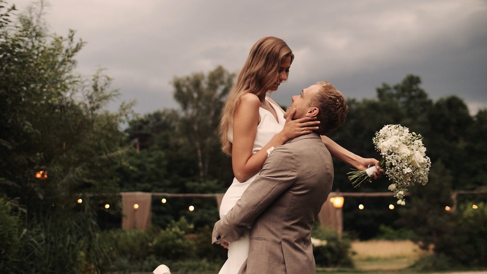 Uśmiechnięta panna młoda w białej sukni, trzymana w objęciach przez pana młodego w brązowym garniturze, z bukietem ślubnym w ręku, na tle zieleni i dekoracyjnych światełek.