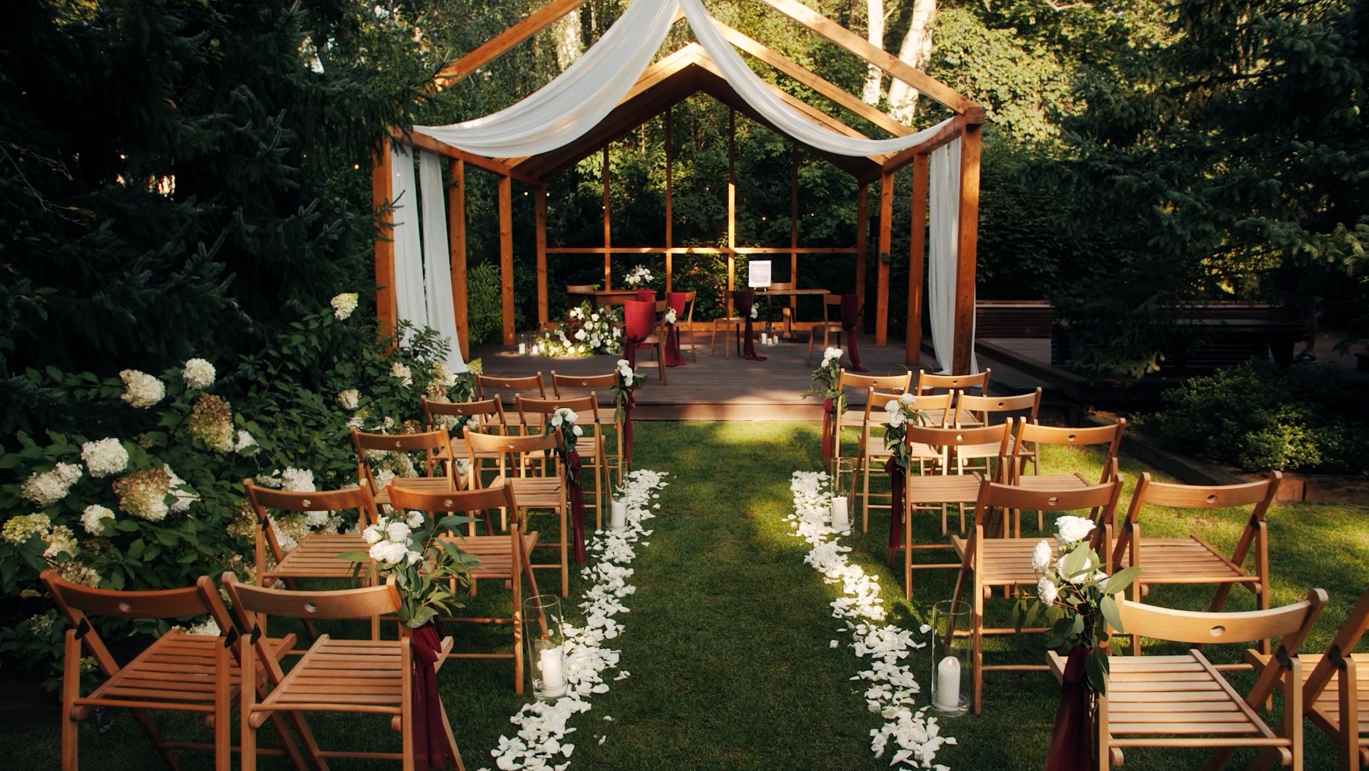 Drewniana konstrukcja z białym materiałem jako zadaszenie, przygotowana na ceremonię ślubną na świeżym powietrzu, z krzesłami ustawionymi w rzędach i ścieżką usłaną białymi płatkami kwiatów.