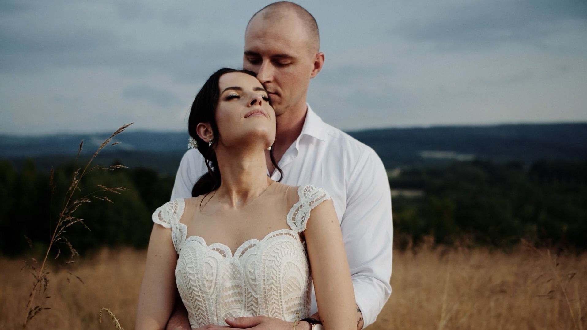 Młoda para w objęciach na tle łąki i zalesionych wzgórz; panna młoda w koronkowej sukni z odkrytymi ramionami, pan młody w białej koszuli.