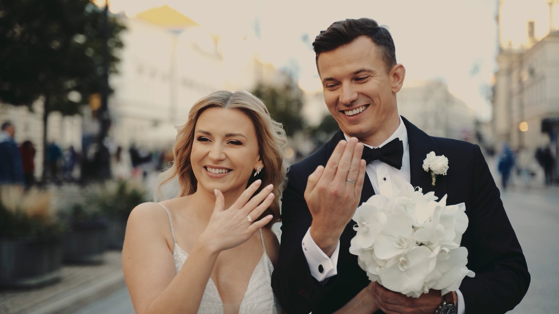 Uśmiechnięta para młoda prezentuje obrączki na tle miejskiej ulicy. Panna młoda w sukni z delikatnymi ramiączkami, pan młody w czarnym smokingu z białą butonierką i bukietem białych kwiatów.