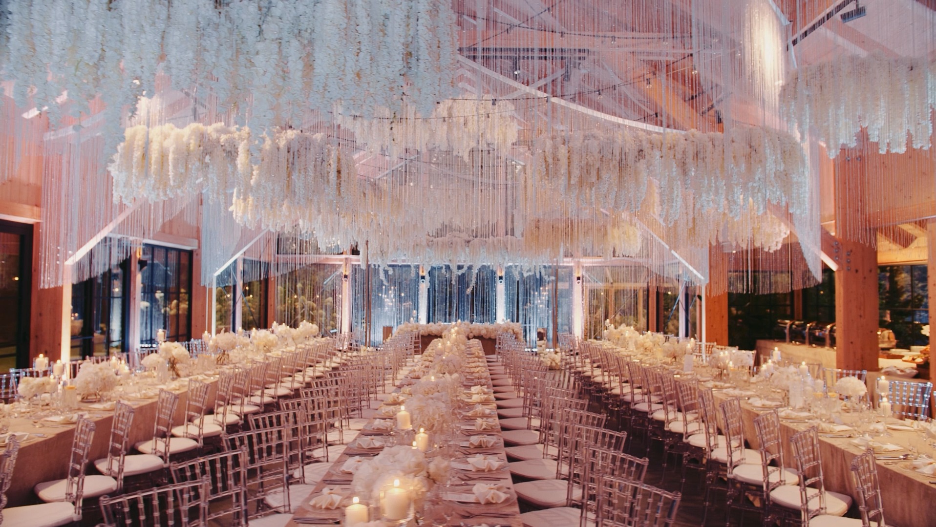 Uroczysta sala weselna z długimi stołami udekorowanymi białymi kwiatami i świecami, nad stołami zwisające dekoracje kwiatowe z białych kwiatów i kryształków, transparentne krzesła.