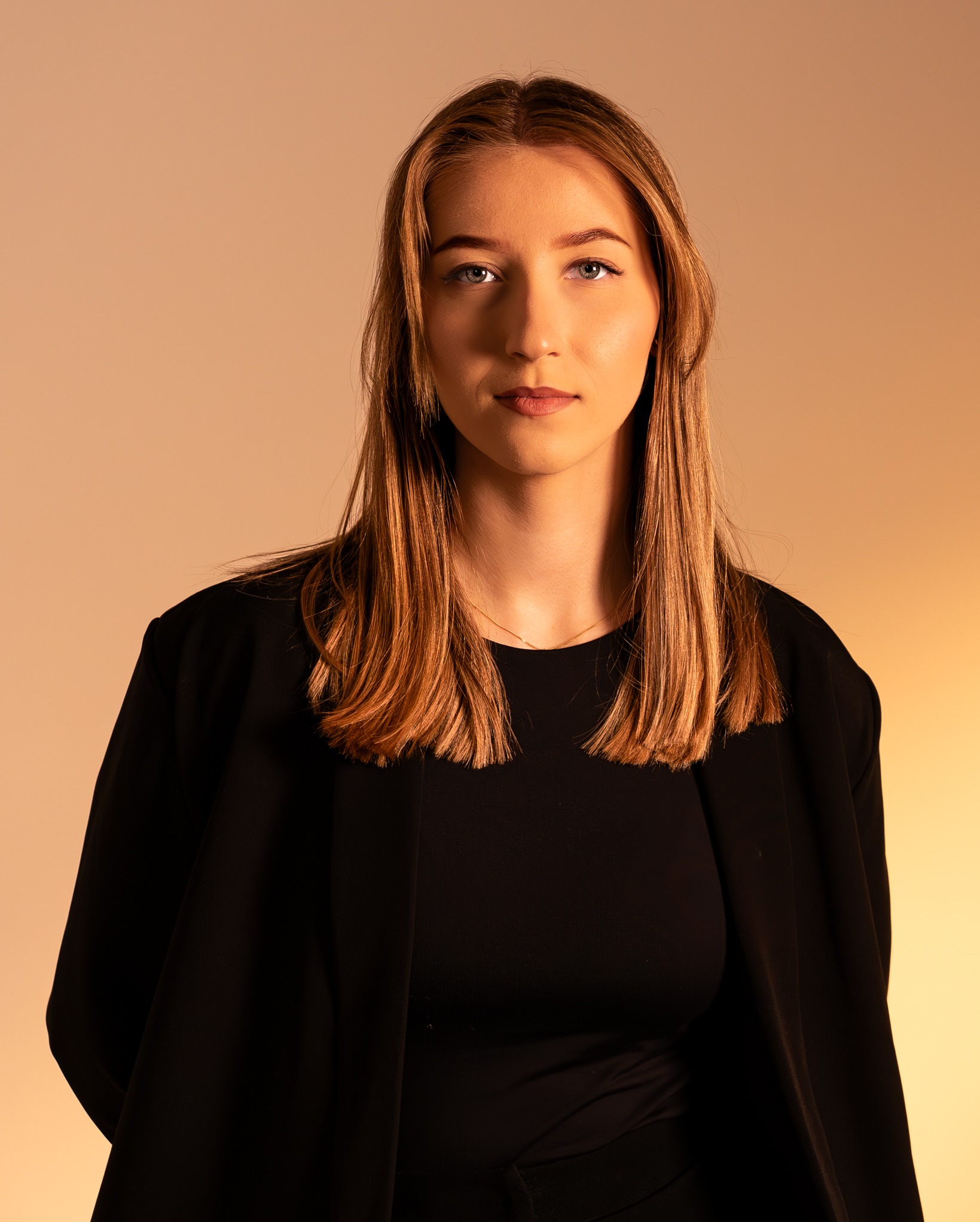 Portret młodej kobiety z długimi, prostymi włosami w czarnej marynarce na jednolitym, ciepłym tle. Ujęcie centralne, skupione na twarzy i górnej części ciała.
