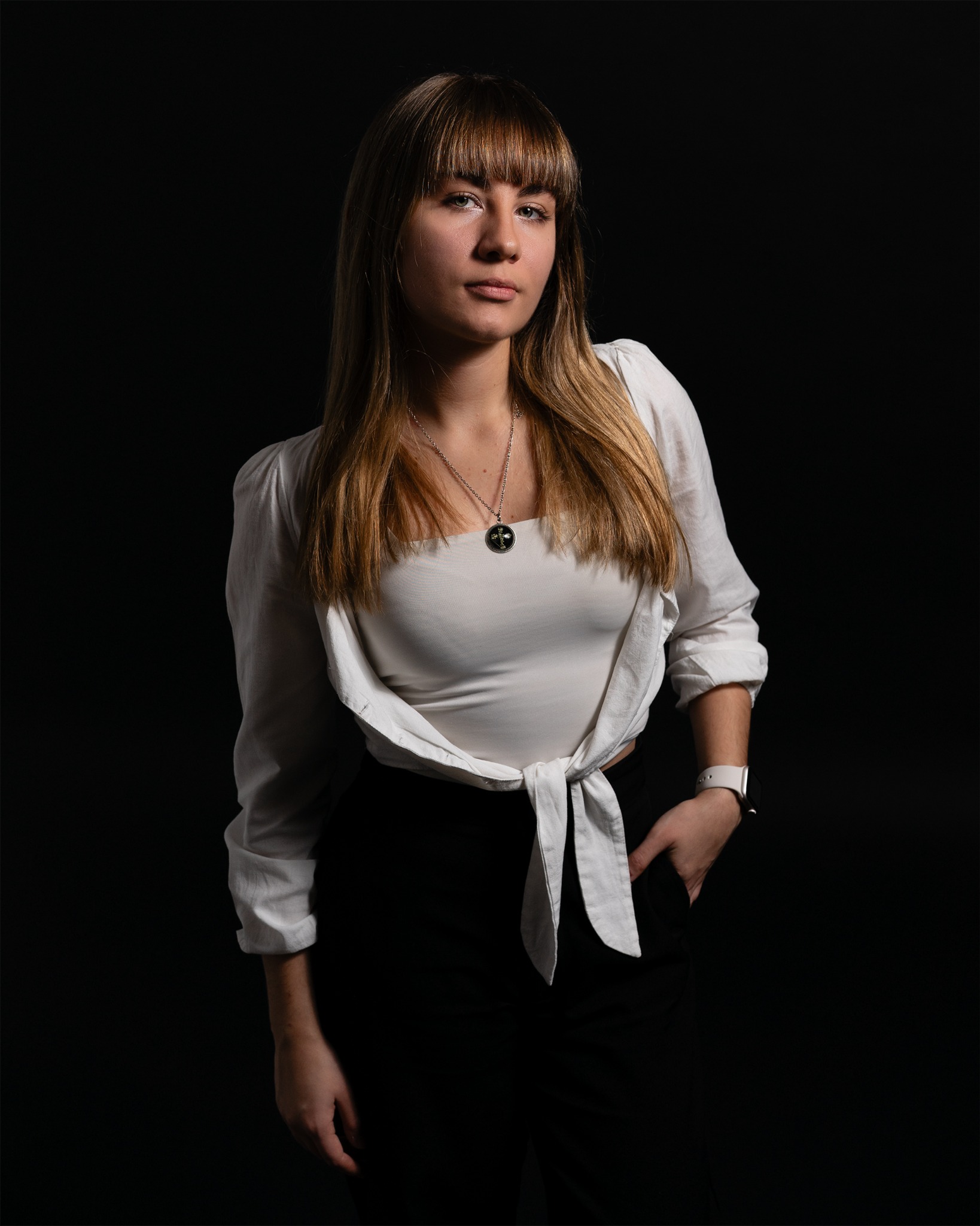 Portret kobiety z grzywką, w białej bluzce i czarnych spodniach, na czarnym tle. Ujęcie studyjne z delikatnym oświetleniem podkreślającym rysy twarzy i detale stroju.