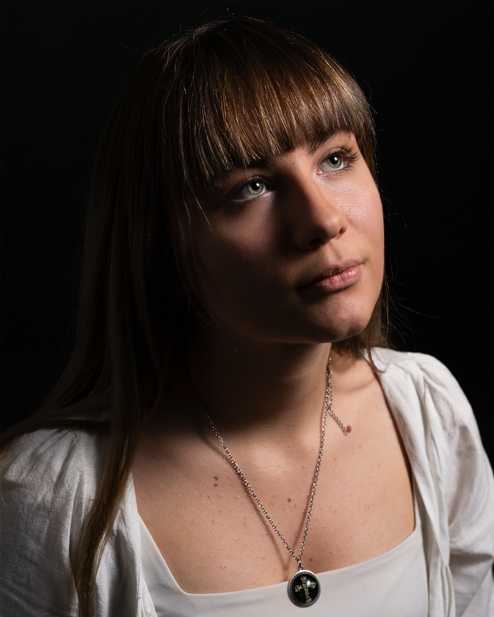 Portret młodej kobiety z grzywką, spoglądającej w górę, na czarnym tle. Delikatne oświetlenie podkreśla rysy twarzy i zielone oczy. Srebrny naszyjnik z krzyżem.