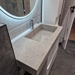 FIRMA OGÓLNOBUDOWLANA PIOTR PIÓRKO - Nowoczesna łazienka z umywalką z szarego kamienia, chromowaną baterią i okrągłym lustrem z podświetleniem LED. W tle widoczny prysznic i grzejnik drabinkowy.