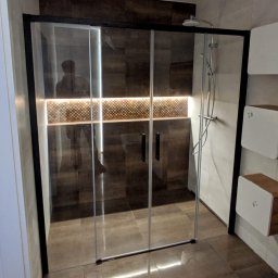 FIRMA OGÓLNOBUDOWLANA PIOTR PIÓRKO - Nowoczesna kabina prysznicowa z przesuwnymi drzwiami w czarnej ramie, podświetlona dekoracyjna ściana z mozaiką, ciemna podłoga z płytek, minimalistyczne półki ścienne po prawej stronie.