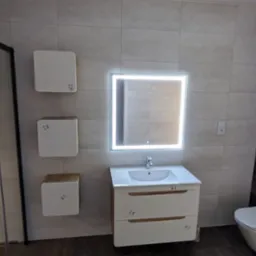 Nowoczesna łazienka z podświetlanym lustrem, wiszącą szafką z umywalką, trzema kwadratowymi szafkami wiszącymi i kabiną prysznicową z czarnymi ramami, wykończona płytkami w odcieniach szarości...