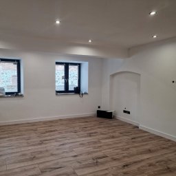 FIRMA OGÓLNOBUDOWLANA PIOTR PIÓRKO - Jasne wnętrze po remoncie z drewnopodobną podłogą, świeżo pomalowanymi ścianami, dwoma oknami z żaluzjami i wnęką w ścianie