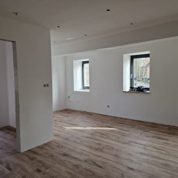 FIRMA OGÓLNOBUDOWLANA PIOTR PIÓRKO - Wnętrze pokoju po remoncie z nową podłogą z paneli imitujących drewno, świeżo pomalowanymi ścianami na biało i zamontowanymi oknami z czarnymi ramami. Widoczne przygotowane otwory pod oświetlenie...