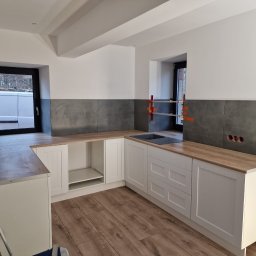 FIRMA OGÓLNOBUDOWLANA PIOTR PIÓRKO - Kątowe ujęcie kuchni w trakcie prac: częściowo położone szare płytki nad blatem z imitacją drewna, białe szafki z frezowanymi frontami, widoczne otwory na gniazdka elektryczne i okno z czarną ramą.