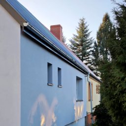 FIRMA OGÓLNOBUDOWLANA PIOTR PIÓRKO - Świeżo pomalowana elewacja domu w odcieniach szarości, z widocznymi panelami słonecznymi na dachu, małymi oknami i refleksami słońca na ścianie, otoczona zielenią.