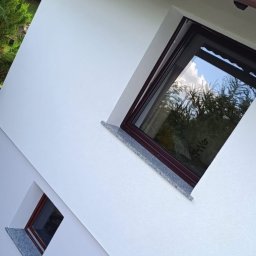 FIRMA OGÓLNOBUDOWLANA PIOTR PIÓRKO - Świeżo pomalowana, biała elewacja domu z brązowymi oknami i parapetami wykończonymi szarym kamieniem, widoczne odbicie drzew w szybach okiennych i fragment brązowej rynny.