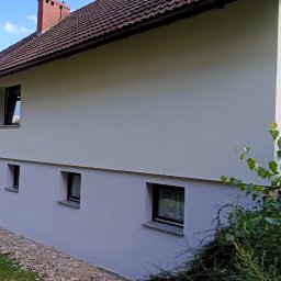 FIRMA OGÓLNOBUDOWLANA PIOTR PIÓRKO - Elewacja domu po malowaniu na szaro, widoczne okna z ciemnymi ramami, dach z brązowej dachówki i fragment ogrodu z zielenią.