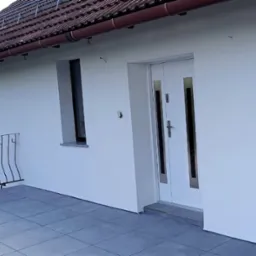 Biała elewacja domu z nowymi oknami i drzwiami wejściowymi, widoczny balkon z metalową balustradą i szara płytka na podłodze, brązowy dach z rynnami.