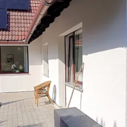 Odnowiona elewacja domu z brązowymi rynnami, panelami słonecznymi na dachu i wybrukowanym tarasem z wiklinowym krzesłem i skrzynią ogrodową