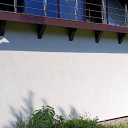 FIRMA OGÓLNOBUDOWLANA PIOTR PIÓRKO - Boczna ściana domu po świeżym pomalowaniu na biało, widoczne brązowe elementy konstrukcyjne dachu, balkon z metalową barierką oraz zieleń w tle.