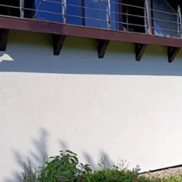 Boczna ściana domu po świeżym pomalowaniu na biało, widoczne brązowe elementy konstrukcyjne dachu, balkon z metalową barierką oraz zieleń w tle.