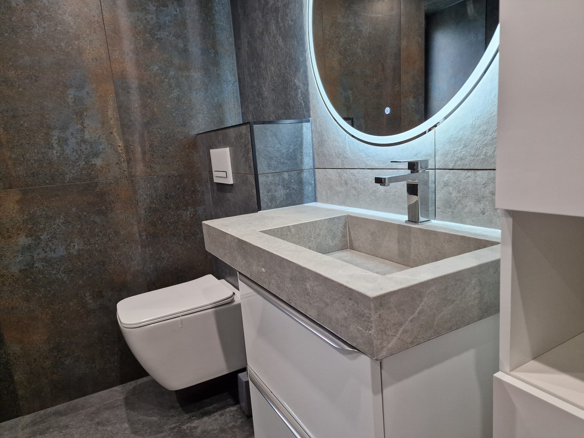 Nowoczesna łazienka z wiszącą toaletą, umywalką z szarego kamienia i okrągłym lustrem z podświetleniem LED, na tle płytek imitujących rdzewiejący metal.