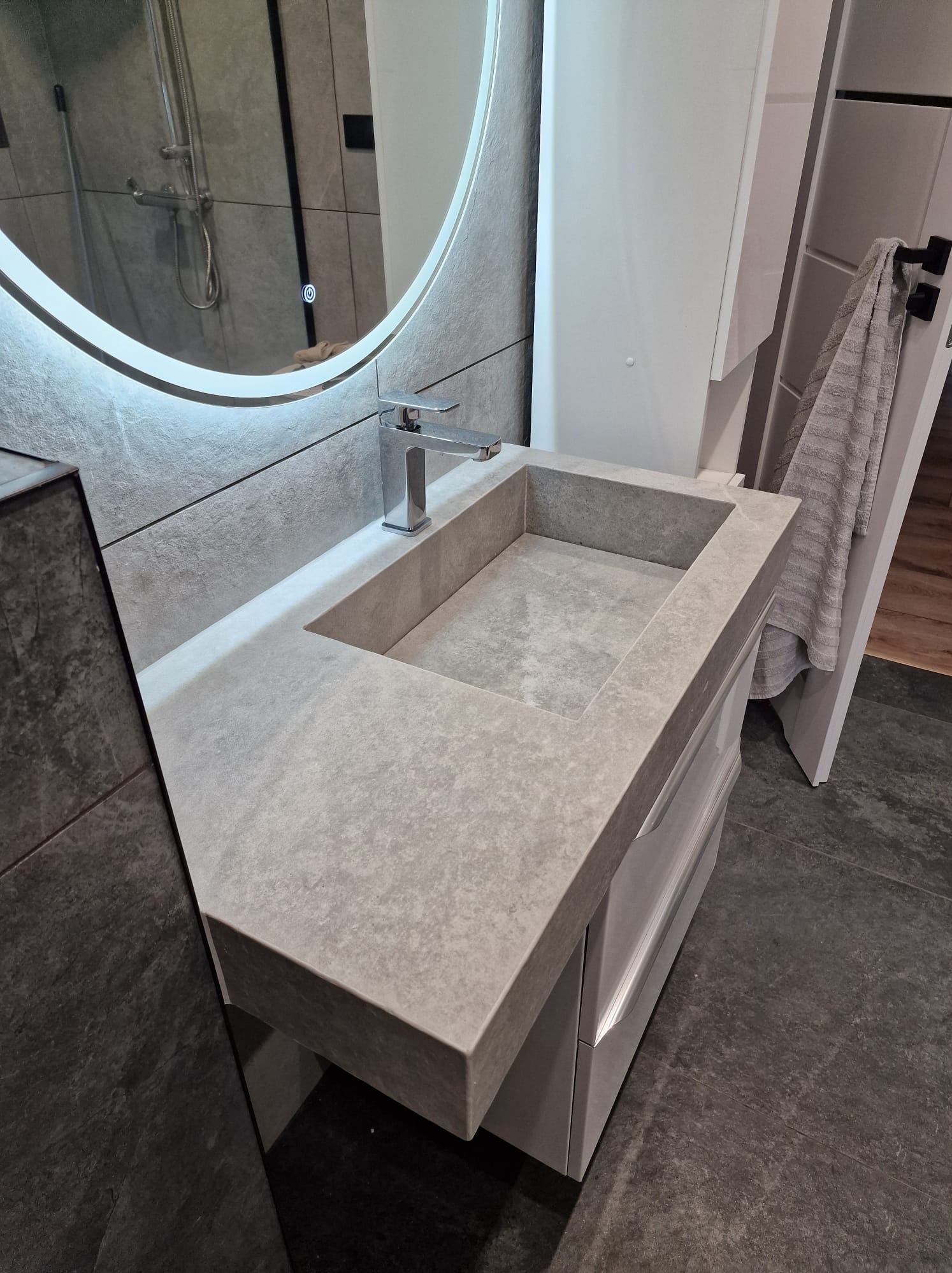 Nowoczesna łazienka z umywalką z szarego kamienia, chromowaną baterią i okrągłym lustrem z podświetleniem LED. W tle widoczny prysznic i grzejnik drabinkowy.