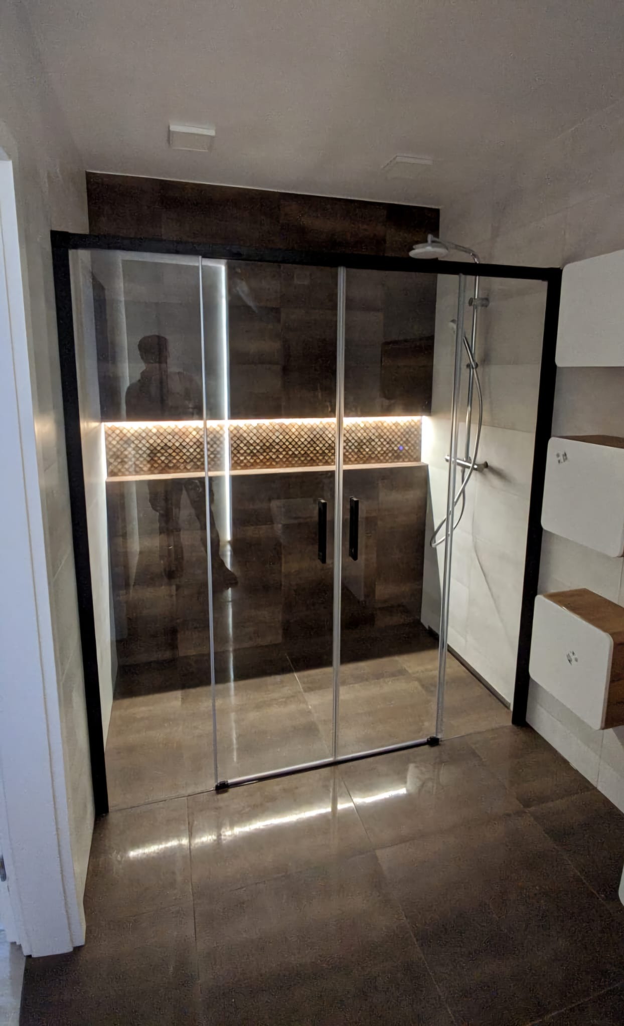 Nowoczesna kabina prysznicowa z przesuwnymi drzwiami w czarnej ramie, podświetlona dekoracyjna ściana z mozaiką, ciemna podłoga z płytek, minimalistyczne półki ścienne po prawej stronie.