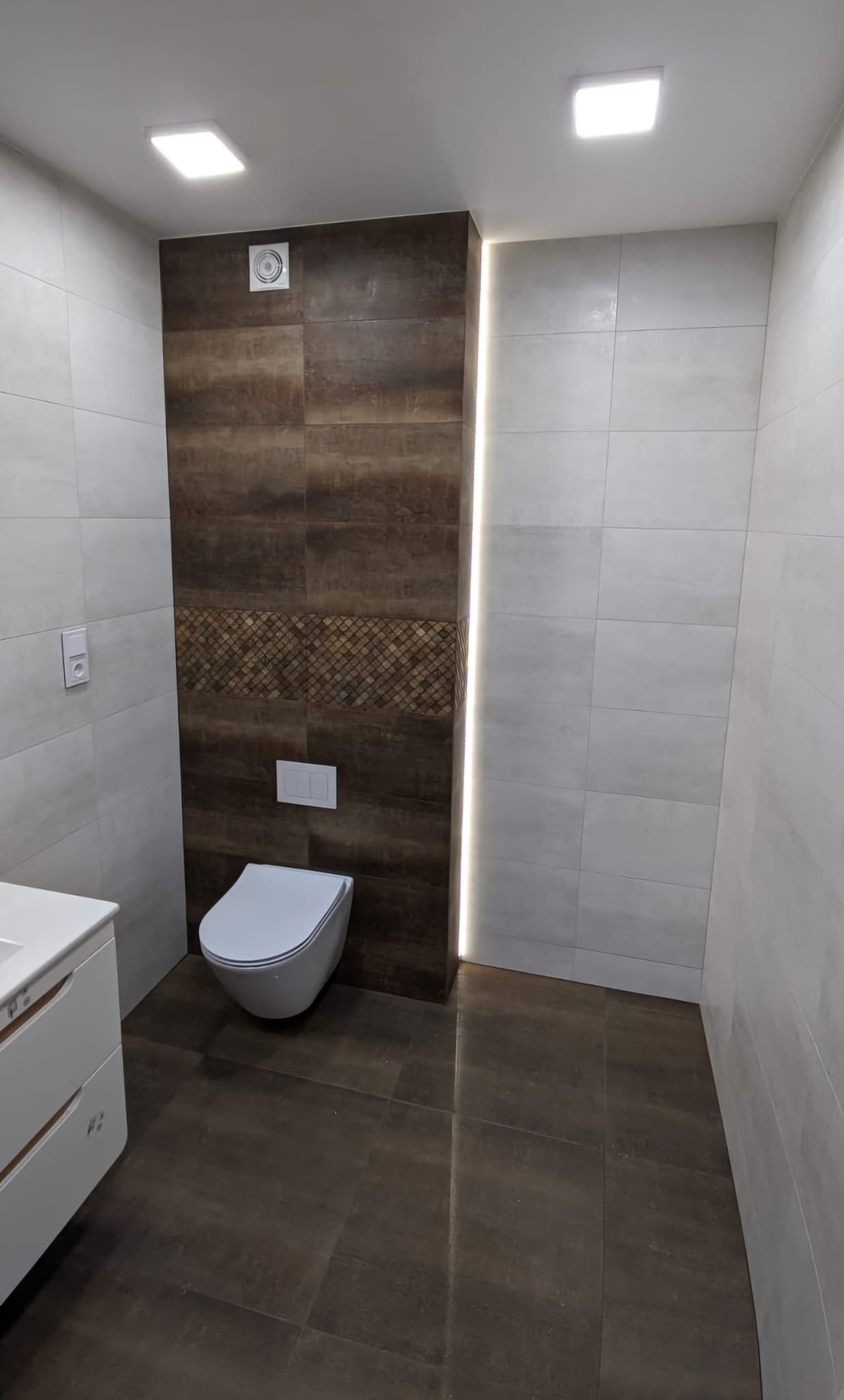 Wykończona łazienka z brązowymi i białymi płytkami, podświetleniem LED w rogu, wiszącą toaletą i wentylatorem sufitowym.