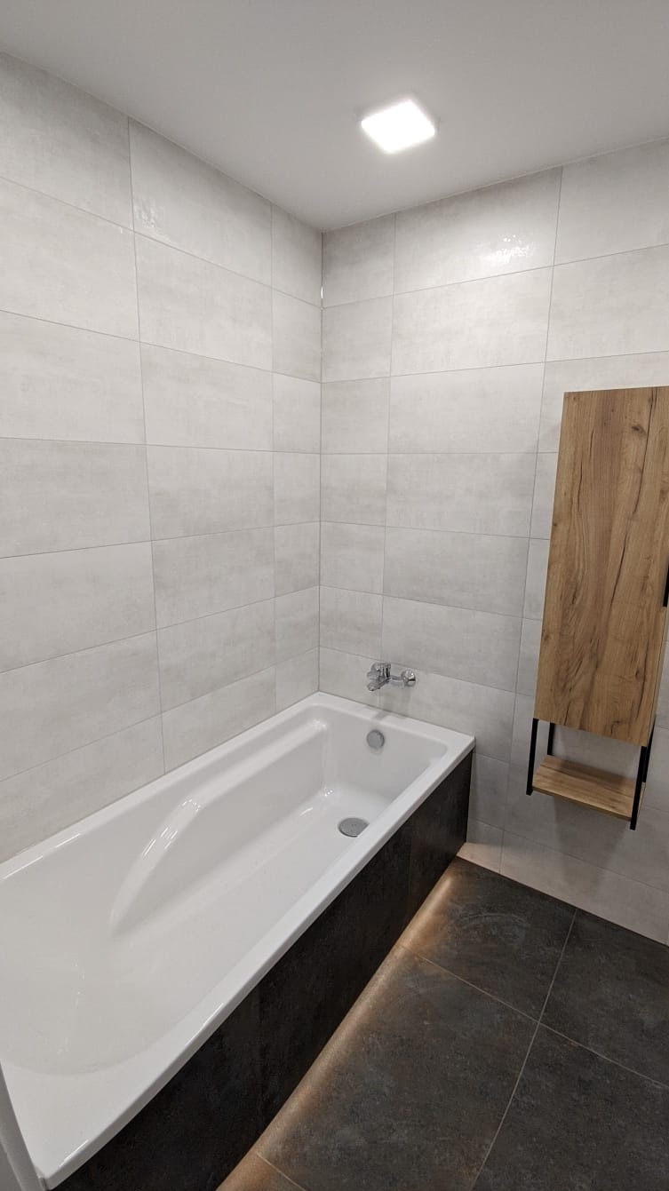 Nowoczesna łazienka po remoncie: biała wanna obudowana ciemnoszarymi płytkami, jasnoszare płytki na ścianach, drewniana szafka wisząca z półką, ciemna podłoga z płytek.