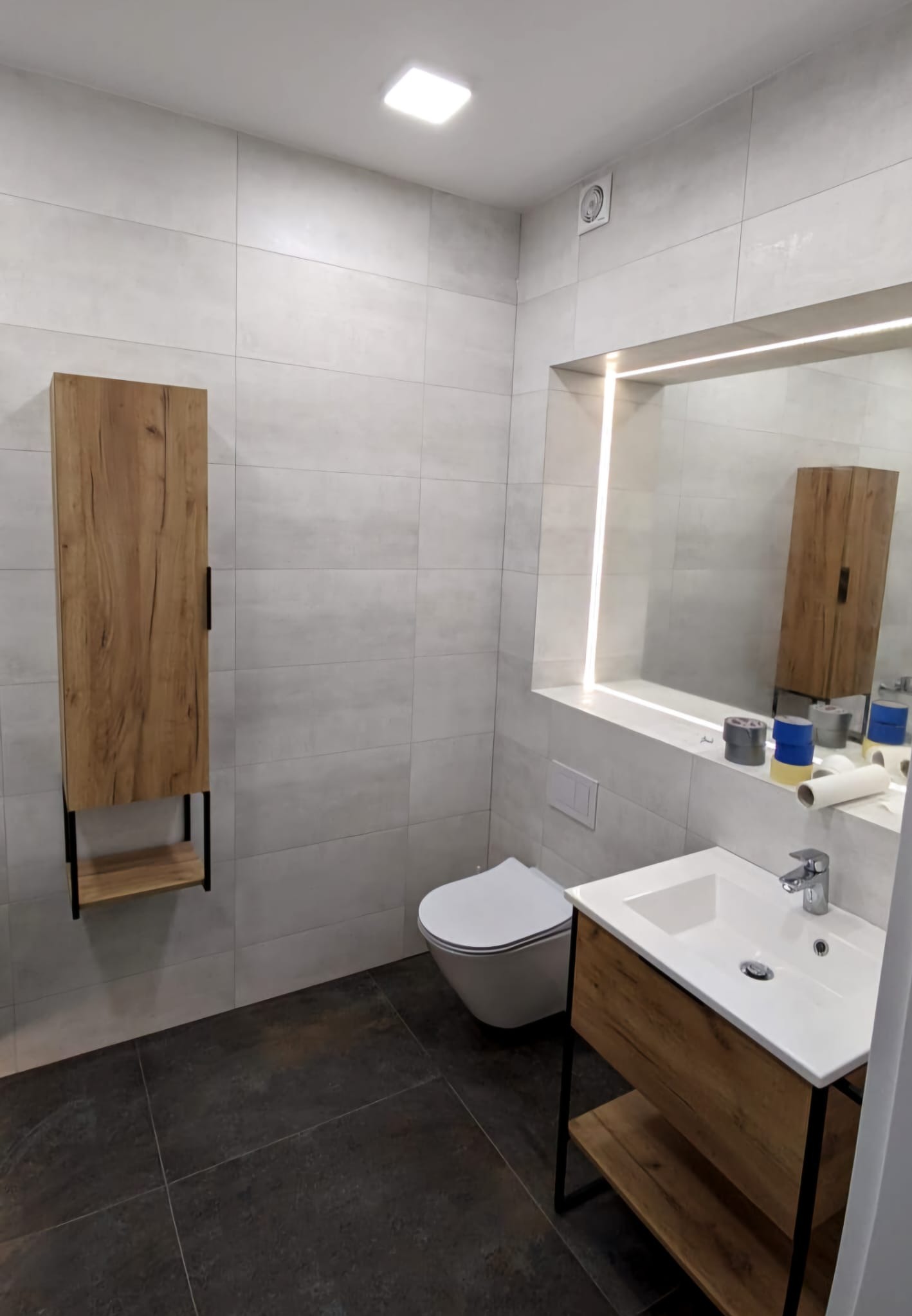 Nowoczesna łazienka z szarymi płytkami, drewnianą szafką pod umywalką i wiszącą szafką, ciemna podłoga, lustro z podświetleniem LED, widoczne taśmy malarskie i rolka papieru na blacie.