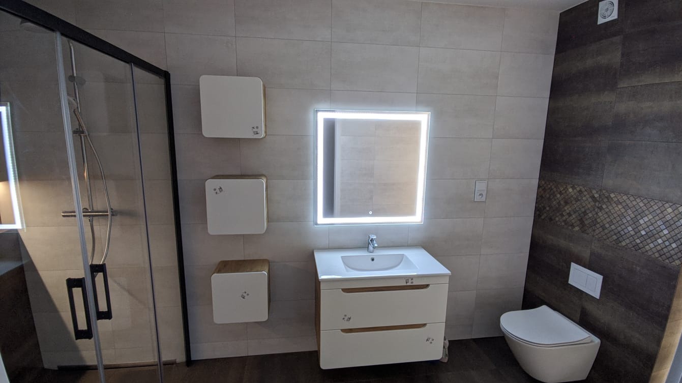 Nowoczesna łazienka z podświetlanym lustrem, wiszącą szafką z umywalką, trzema kwadratowymi szafkami wiszącymi i kabiną prysznicową z czarnymi ramami, wykończona płytkami w odcieniach szarości...