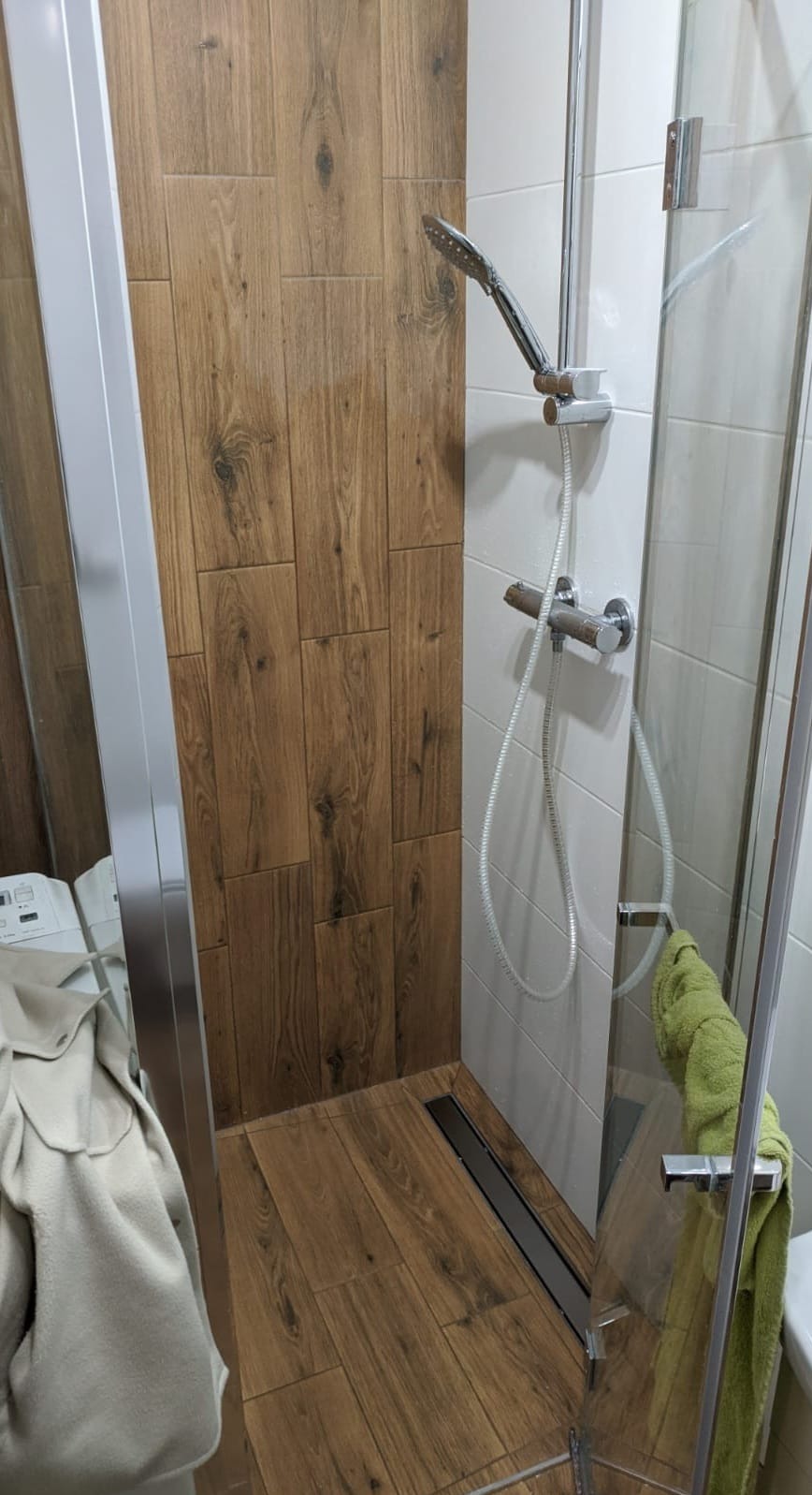 Nowoczesna kabina prysznicowa z odpływem liniowym i płytkami imitującymi drewno na ścianie, widoczny fragment pralki i ręcznik na drzwiach kabiny.