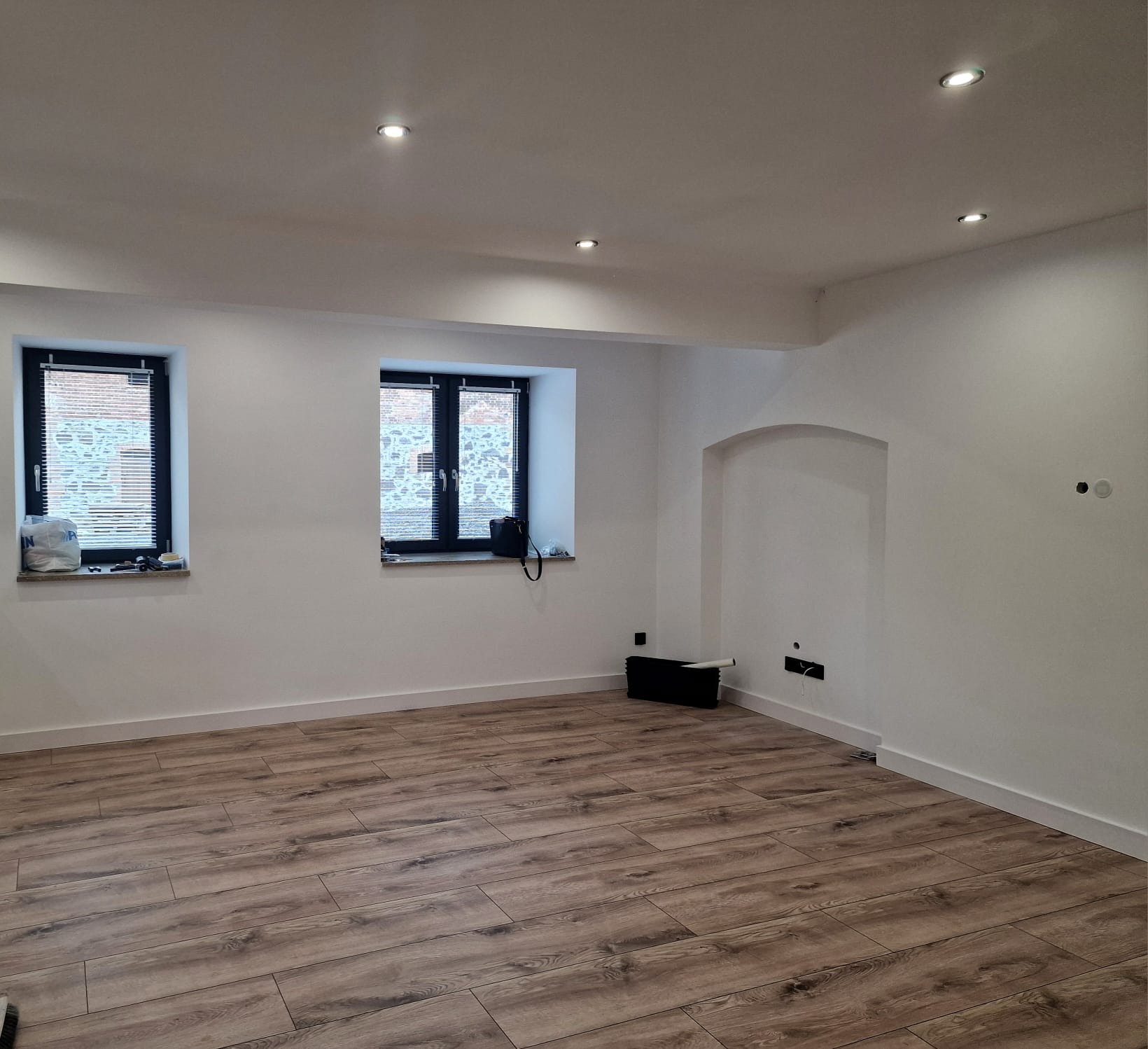 Jasne wnętrze po remoncie z drewnopodobną podłogą, świeżo pomalowanymi ścianami, dwoma oknami z żaluzjami i wnęką w ścianie